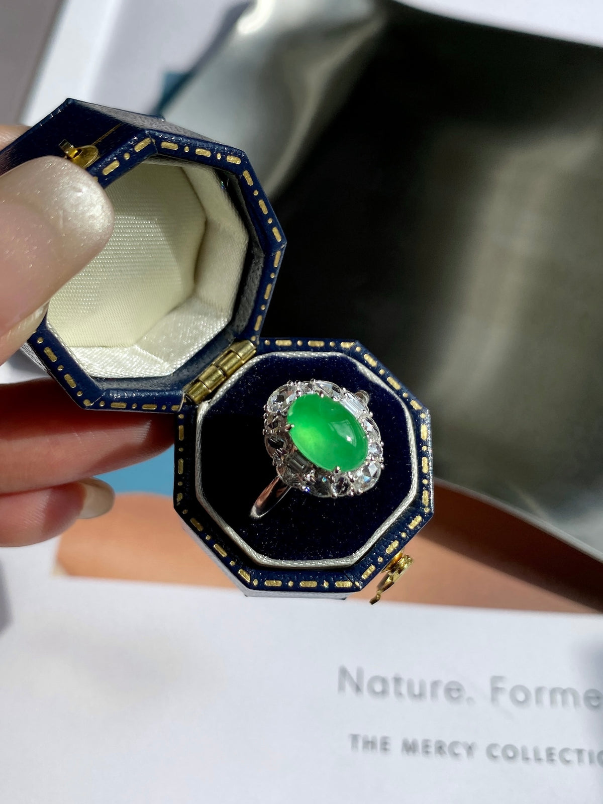 Green jade cabochon ring with ornate silver-tone halo, displayed in open ring box