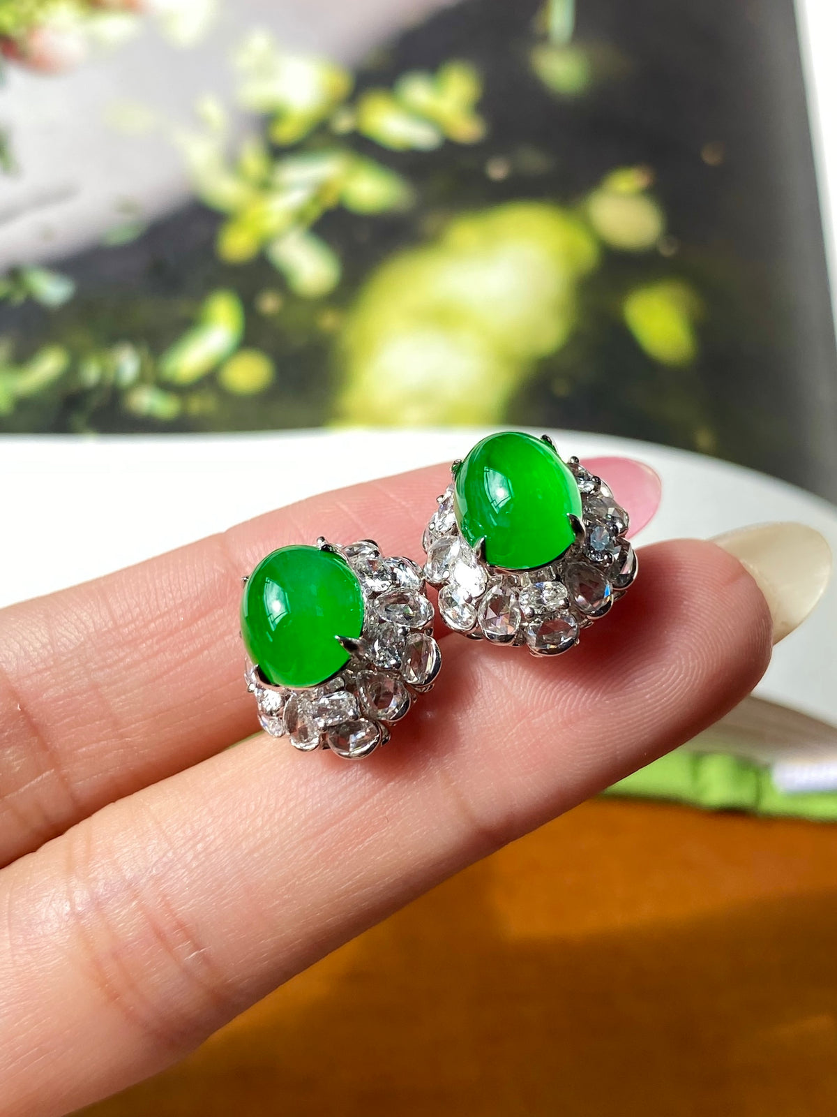 Green jade cabochon stud earrings with silver filigree flower halos, close-up on fingers