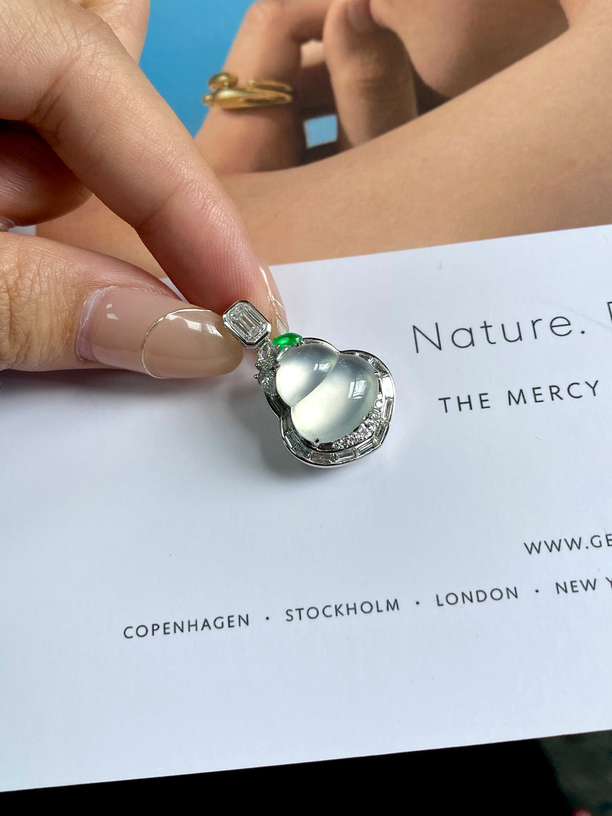 Silver gourd pendant with oval light green stone and tiny green gem, held on paper